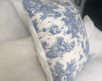 May include: A white pillow with a blue and white toile pattern featuring a pastoral scene. The pillow is on a white bed.