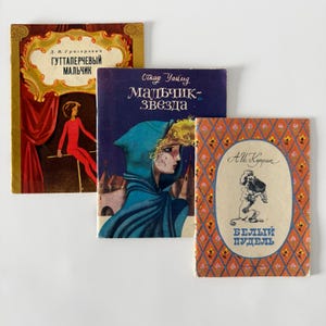 May include: Three vintage books with illustrated covers. The left book features a red-clad figure on a stage. The center book shows a person in a blue hood with the text Malchik-Zvezda. The right book has an orange floral pattern and a poodle illustration.
