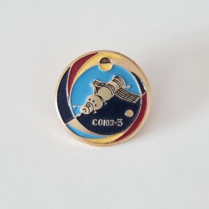 May include: A round, gold-colored pin featuring a space shuttle design with the text "COЮЗ-3" in white. The pin has a blue, yellow, and red background, with a small hole at the top. The pin is likely a collectible or souvenir.