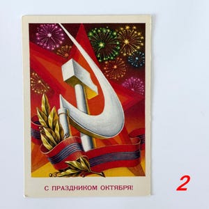 May include: A rectangular postcard featuring a white hammer and sickle symbol centered over a large red star. The background displays colorful fireworks. A red ribbon with dark stripes and green laurel leaves sits below. Text reads: С ПРАЗДНИКОМ ОКТЯБРЯ!