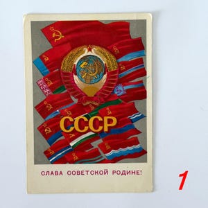 May include: A rectangular postcard featuring a central Soviet emblem with a globe, hammer, and sickle surrounded by wheat stalks. Multiple red flags with gold symbols are arranged behind the emblem. The text CCCP is in gold, with SLAVA SOVETSKOY RODINE! printed in red at the bottom.