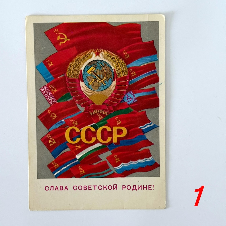 May include: A rectangular postcard featuring a central Soviet emblem with a globe, hammer, and sickle surrounded by wheat stalks. Multiple red flags with gold symbols are arranged behind the emblem. The text CCCP is in gold, with SLAVA SOVETSKOY RODINE! printed in red at the bottom.