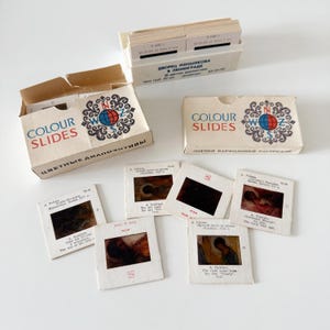 Op de afbeelding: Vintage "Colour Slides" dozen en dia's. De beige dozen hebben een wereldbollogo en tekst. Er zijn verschillende dia's zichtbaar, elk met een kleine afbeelding en tekst. De dia's zitten in een witte doos.