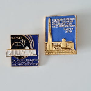 Puede incluir: Dos pines de esmalte azul y dorado que representan el Museo Estatal de Historia de la Cosmonáutica en Kaluga, Rusia. El pin superior muestra un cohete y el pin inferior muestra el edificio del museo. Ambos pines tienen el texto "Kaluga 1976" y el nombre del museo en ruso.