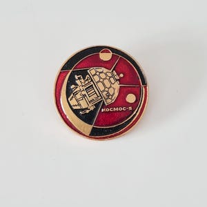 May include: A red, black, and gold round pin with a stylized image of a satellite and the text "KOSMOS-5".