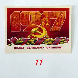 May include: A rectangular postcard featuring the red text 1917 with industrial illustrations inside the numbers, a gold hammer and sickle emblem, and a green laurel branch wrapped in a red and blue ribbon, with the text SLAVA VELIKOMU OKTYABRYU!