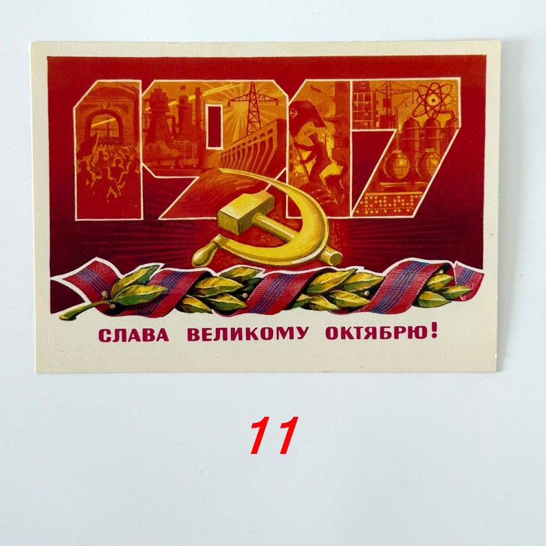 May include: A rectangular postcard featuring the red text 1917 with industrial illustrations inside the numbers, a gold hammer and sickle emblem, and a green laurel branch wrapped in a red and blue ribbon, with the text SLAVA VELIKOMU OKTYABRYU!