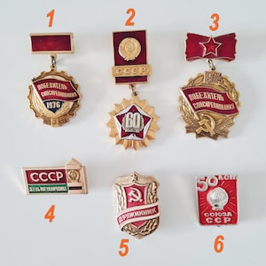 May include: A collection of six vintage Soviet-era pins and medals. The pins feature red, gold, and white enamel with Cyrillic text and symbols, including stars, hammers, sickles, and the CCCP emblem. The pins are likely commemorative.