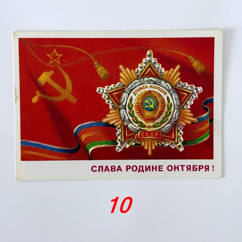 May include: A rectangular postcard featuring a red background with a gold hammer and sickle emblem. A detailed Order of Friendship of Peoples medal is centered, with a ribbon below and the text SLAVA RODINE OKTYABRYA in red capital letters.