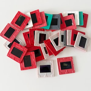 May include: A collection of vintage 35mm slide mounts in various colors, including red, white, gray, and teal. The slides have a rectangular shape with a black viewing area. The mounts are scattered on a white surface.