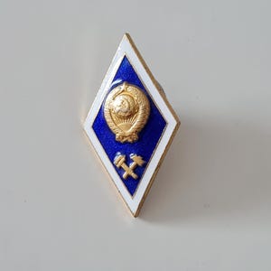 May include: A diamond-shaped pin with a blue center, white border, and gold accents. The pin features a gold emblem of a hammer and sickle, and a wheat sheaf. The pin is likely a vintage or collectible item.
