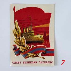 May include: A rectangular postcard featuring a graphic illustration of a battleship against a red background. A golden laurel branch and a red, blue, and gold ribbon are in the foreground. The text at the bottom reads: СЛАВА ВЕЛИКОМУ ОКТЯБРЮ!