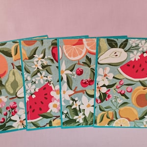 May include: Set of four rectangular fabric coasters with a light blue background. Each coaster features a different fruit design, including oranges, pears, watermelon, and lemons, along with white flowers and green leaves. The edges are trimmed with a teal border.