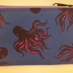 May include: A rectangular blue zippered pouch featuring a repeating pattern of black octopuses with pink suckers. The zipper is black, and the lining is purple. The pouch is likely made of fabric and is suitable for carrying small items.