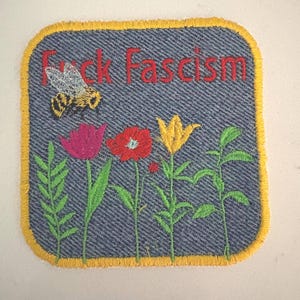 May include: A square denim patch with a yellow stitched border featuring an embroidered bee, a pink flower, a red flower, a yellow flower, and green stems. The text "Fuck Fascism" is embroidered in red thread across the top of the patch.