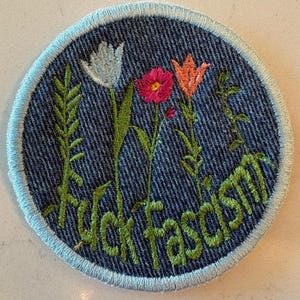 May include: Circular embroidered patch with a denim effect background and a pale blue border. It features embroidered flowers in white, pink, and orange, with green stems. The words "fuck fascism" are in green.