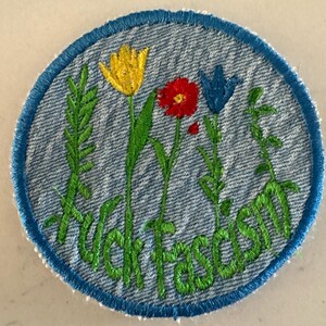 May include: A circular denim patch with a blue stitched border featuring embroidered green stems, a yellow tulip, a red poppy, and a blue bellflower. The text "fuck fascism" is embroidered in green cursive script across the bottom.