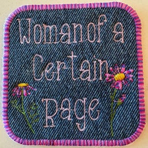 May include: Square denim patch with embroidered text "Woman of a Certain Rage" in light pink thread. Two embroidered flowers with pink and purple petals and yellow centres are also featured. The patch has a pink and purple stitched border.