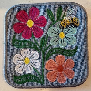May include: Embroidered patch with a denim effect background, featuring colourful flowers and a bee. The flowers are in shades of pink, blue, white, and orange, each with a yellow centre. The bee is black and yellow. The patch has a grey border.