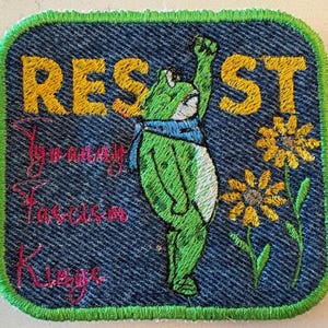 May include: Embroidered patch with a denim-style background and a green border. The patch features a green frog with a raised fist, a blue scarf, and yellow text that reads "RESIST". Additional text includes "Tyranny, Fascism, Kings".