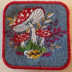 May include: Embroidered patch featuring a cluster of red and white mushrooms with white stems, surrounded by green grass, yellow and burgundy leaves. The patch has a denim background with a red border.