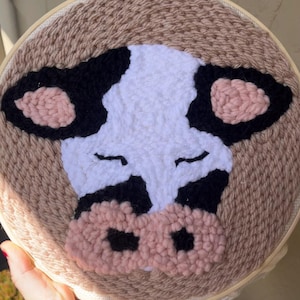 May include: A close-up of a hand-crafted cow face made with wool yarn in a circular embroidery hoop. The cow has black and white spots, pink ears and a pink nose.