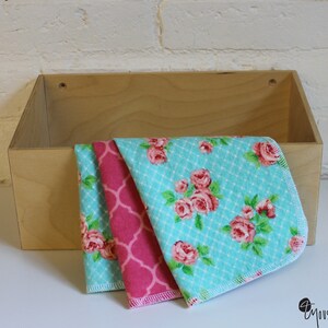 May include: Three fabric squares with floral and geometric patterns. The squares are folded and stacked on top of each other. The top square is turquoise blue with a white grid pattern and pink roses. The middle square is pink with a geometric pattern. The bottom square is turquoise blue with a white grid pattern and pink roses.