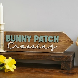 May include: A wooden sign in the shape of an arrow, with the words "BUNNY PATCH Crossing" in teal and white. A white ceramic rabbit and a candle holder with a lit candle are on display. Yellow daffodils in a woven basket are also present.