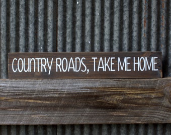 May include: A rustic wooden sign with the text "COUNTRY ROADS, TAKE ME HOME" painted in white.
