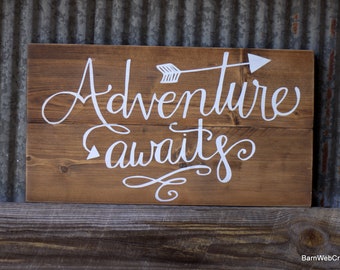 May include: A rustic wooden sign with the words "Adventure awaits" written in white cursive lettering. An arrow pointing to the right is above the word "Adventure".