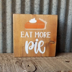 May include: A wooden sign with the text "Eat More Pie" in white lettering. A stylized orange pie with white whipped cream is depicted above the text.