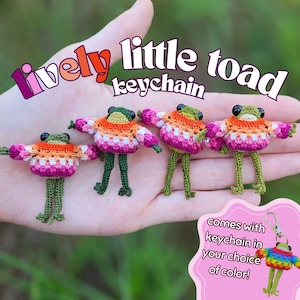 Op de afbeelding: Vier kleine gehaakte padfiguren met zwarte kraaloogjes en lange groene poten. Elke pad draagt een gestreepte trui in oranje, wit en roze. De tekst luidt "lively little toad keychain" en "comes with keychain in your choice of color!"