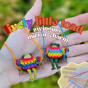 May include: Two crocheted toad figures sitting on small wooden swings, each wearing a colorful rainbow sweater. The text 'lively little toad swinging mirror charm' is displayed above, with a graphic showing color options in the corner.