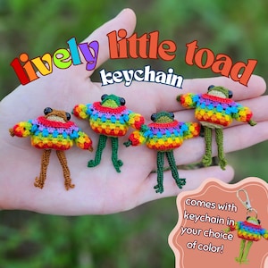 May include: Four small crocheted toad figures, each wearing a colorful rainbow-patterned sweater. The text "lively little toad keychain" appears at the top, and a small inset box shows a toad attached to a metal keychain with the text "comes with keychain in your choice of color!"