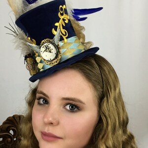 May include: A navy blue top hat with gold accents and a clock face, adorned with white and blue feathers. The hat is worn on a person's head.