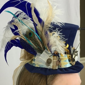 May include: A blue top hat with a gold band and a clock face design. The hat is decorated with feathers in shades of blue, white, and teal, as well as a peacock feather.