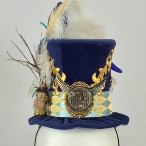 May include: A blue velvet top hat with gold accents and a clock face design. The hat has a white feather and a small bottle with a key attached.