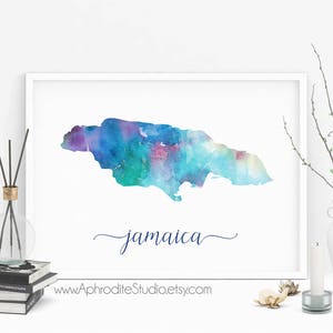 May include: A watercolor painting of the island of Jamaica in shades of blue, green, and purple. The word "Jamaica" is written in cursive below the island. The painting is framed and displayed on a white wall.