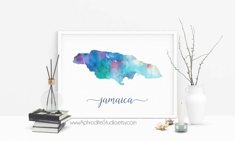 May include: A watercolor painting of the island of Jamaica in shades of blue, green, and purple. The word "Jamaica" is written in cursive below the island. The painting is framed and displayed on a white wall.