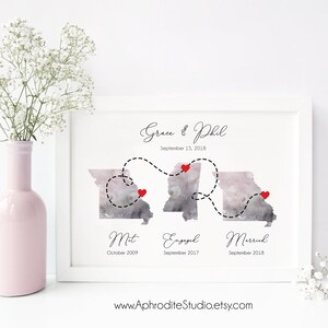 May include: A watercolor print of a couple's love story timeline. The print features two watercolor illustrations of the state of Missouri, with red hearts marking the locations where the couple met, got engaged, and married. The print includes the couple's names, "Grace & Phil", and the dates of each milestone. The text "September 15, 2018" is also included. The print is framed in a white frame.