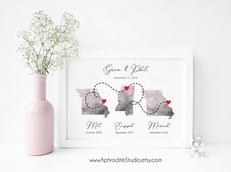 May include: A watercolor print of a couple's love story timeline. The print features two watercolor illustrations of the state of Missouri, with red hearts marking the locations where the couple met, got engaged, and married. The print includes the couple's names, "Grace & Phil", and the dates of each milestone. The text "September 15, 2018" is also included. The print is framed in a white frame.