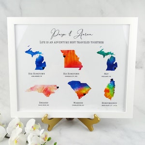 May include: A watercolor print of a couple's journey together, featuring maps of their hometown, where they met, got engaged, married, and honeymooned. The print is framed in white and includes the text "Paige & Aeron, Life is an adventure best traveled together."