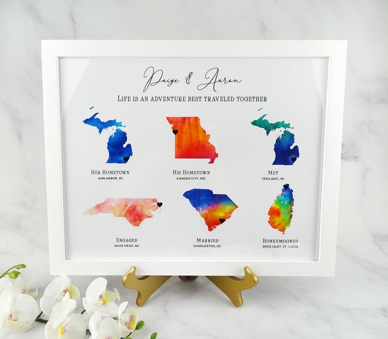 May include: A watercolor print of a couple's journey together, featuring maps of their hometown, where they met, got engaged, married, and honeymooned. The print is framed in white and includes the text "Paige & Aeron, Life is an adventure best traveled together."