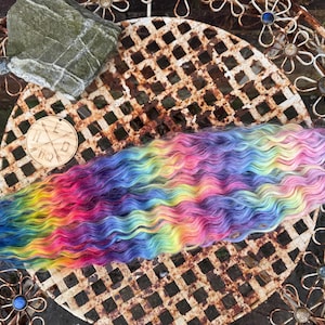 May include: A long strand of wavy, rainbow-colored hair. The hair is dyed in shades of blue, green, yellow, orange, red, pink, and purple. The hair is lying on a rusty metal surface.