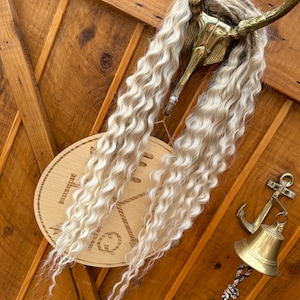 May include: A wooden sign with the text "The Hang" carved into it, hanging from a gold metal hook with blonde curly hair hanging from the hook. A gold bell with an anchor on top is hanging from a rope on the right side of the image.
