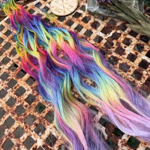 Könnte beinhalten: Eine lange Strähne regenbogenfarbener Haarverlängerungen. Das Haar ist wellig und hat Farbtöne von Rosa, Blau, Gelb, Grün und Lila.