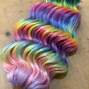 May include: A long, wavy hair extension with a rainbow ombre color pattern. The hair is styled in loose waves and features shades of purple, blue, green, yellow, orange, and pink.