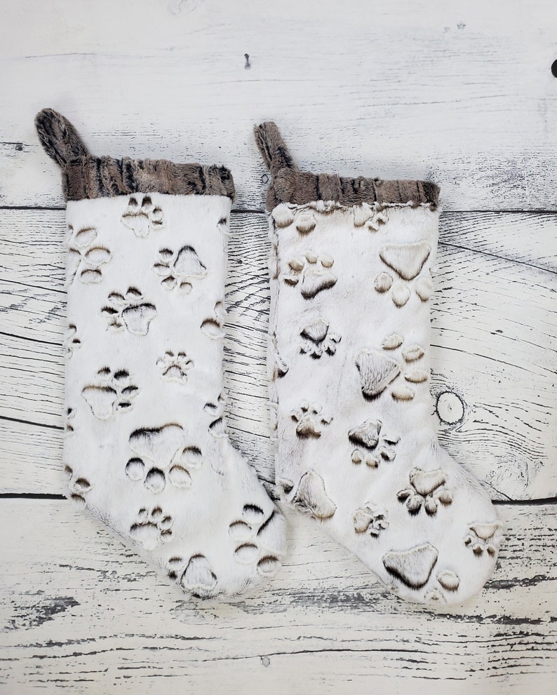 May include: Two white faux fur Christmas stockings with a brown faux fur cuff. The stockings are decorated with a paw print pattern.