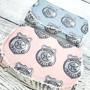 May include: Two fabric pouches with a police badge pattern. The top pouch is light blue and the bottom pouch is pink. Both pouches have a white fringe around the edges.