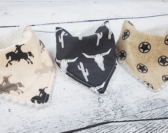 May include: Three bandana bibs with different western-themed prints. One bib features a black and white print of cowboys riding horses, another has a black and white print of cactus and bull skulls, and the last bib has a tan and black print of stars.
